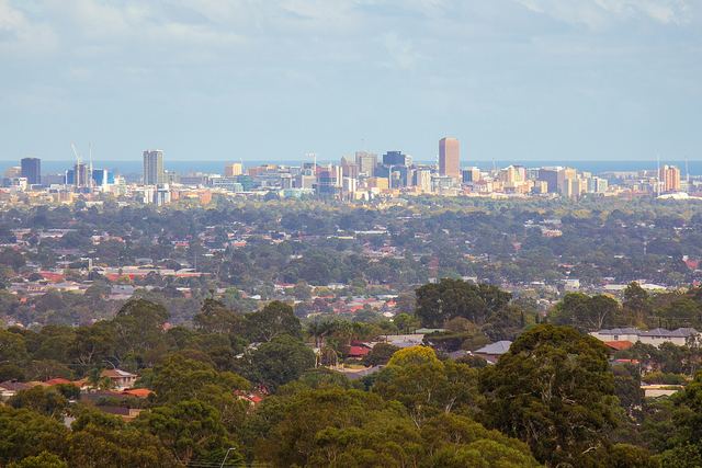 Adelaide city skyline seen from the eastern foothills over leafy suburbs, with the gulf on the horizon.
