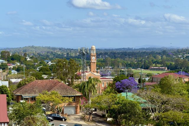 View over Gympie rooftops to the brick Town Hall clock tower, with flowering jacarandas and green hills behind.