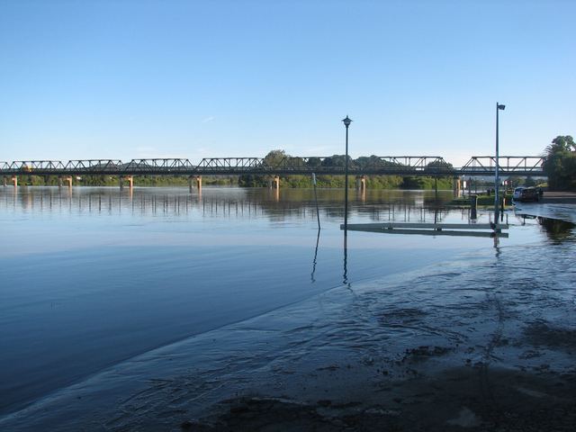 The Manning River at Taree running high over a riverside boat ramp, with the long steel truss Martin Bridge behind.