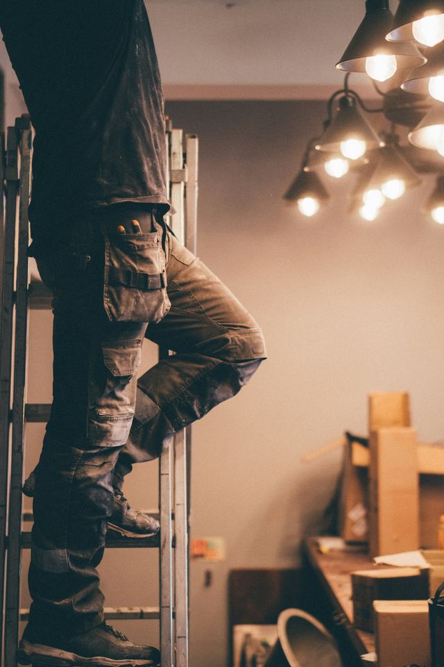 A tradesperson in dusty work pants with a tool pouch standing on a ladder beside a hanging pendant light, seen from the waist down.