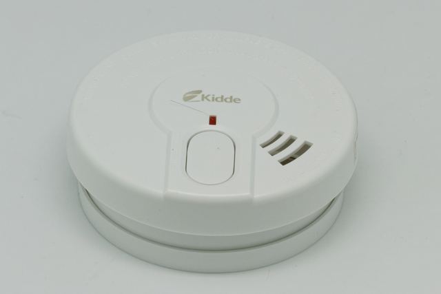 A round white household smoke alarm with a large test button and a small red indicator light, on a plain grey background.
