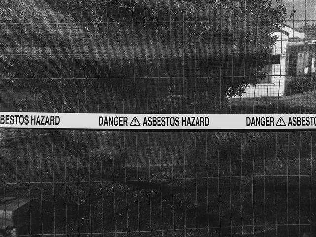 Black and white photo of 'Danger Asbestos Hazard' tape on a temporary fence with shade cloth.