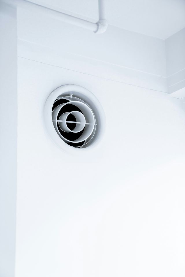 A round white exhaust vent set high in a white wall near the ceiling.