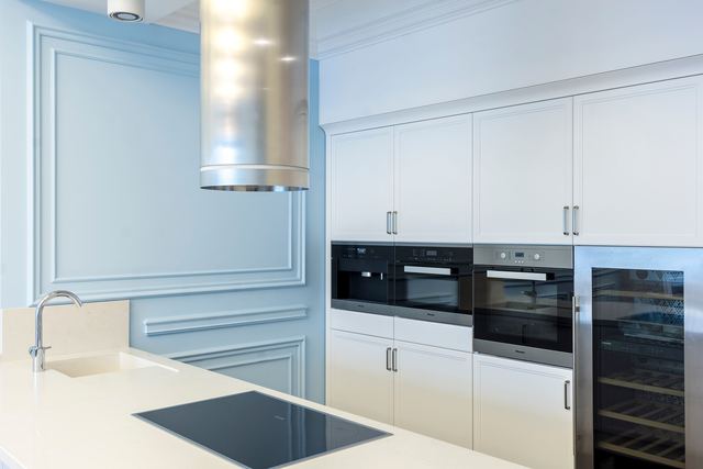 A black induction cooktop set into a white kitchen island under a stainless steel rangehood, with built-in ovens behind.