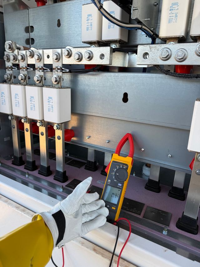 A gloved hand beside a clamp meter reading 0.1 in front of large fuses and busbars inside open switchgear.