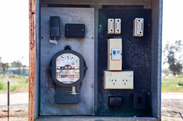 An open outdoor meter box with an old dial electricity meter, ceramic fuses, a safety switch and a double power point.