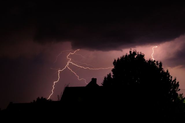 Lightning forks across a purple night sky above the silhouette of a house roof and a large tree.